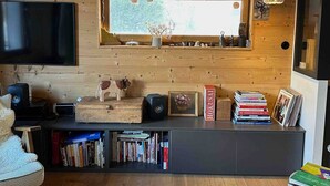 A cozy living room with a wooden wall, a flat-screen TV, a bookshelf, and a window with a view of trees. - Le Nevada - Duplex T4  Proche Centre (Chamonix-Mont-Blanc)
