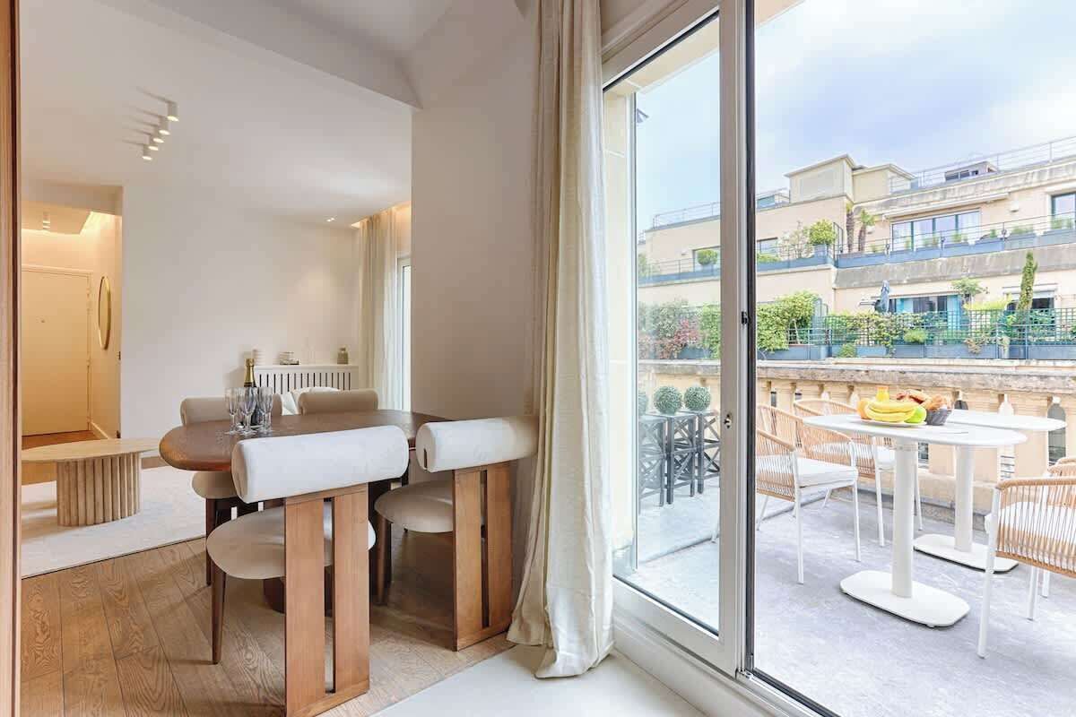 A modern hotel room with a large window, a dining area with a table and chairs, and a view of the outdoor balcony.