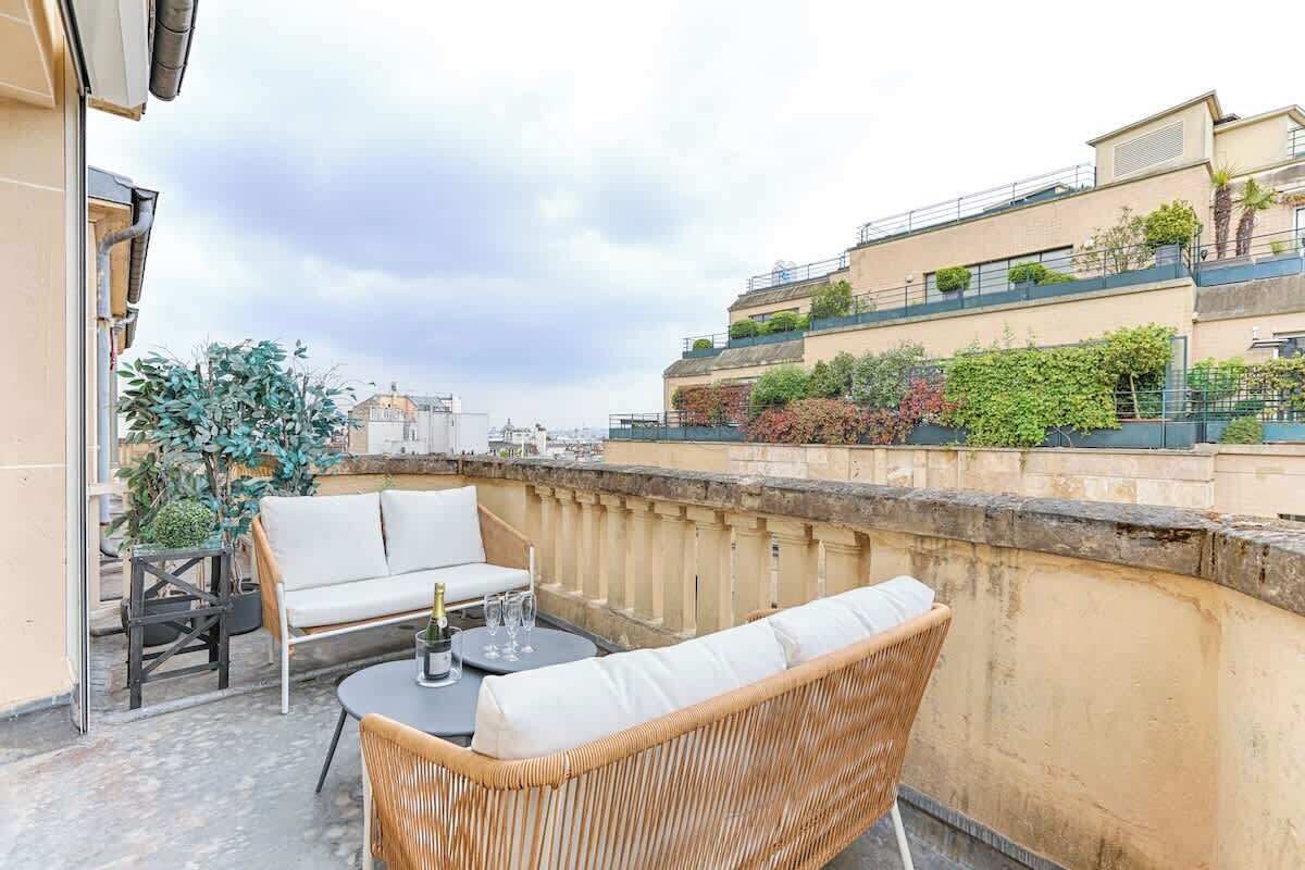 A rooftop terrace with outdoor seating, a view of neighboring buildings, and a clear sky.