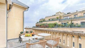 A balcony with wicker chairs and a table, a tiled wall, and a building with balconies in the background. - Appartement de Luxe avec terrasse Champs Elysées (Paris)