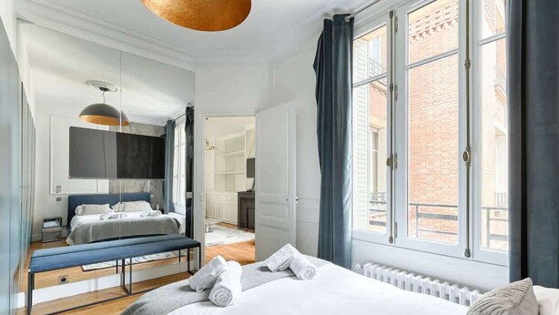 Een moderne slaapkamer met een groot bed, een bankje en uitzicht op een bakstenen gebouw door grote ramen.