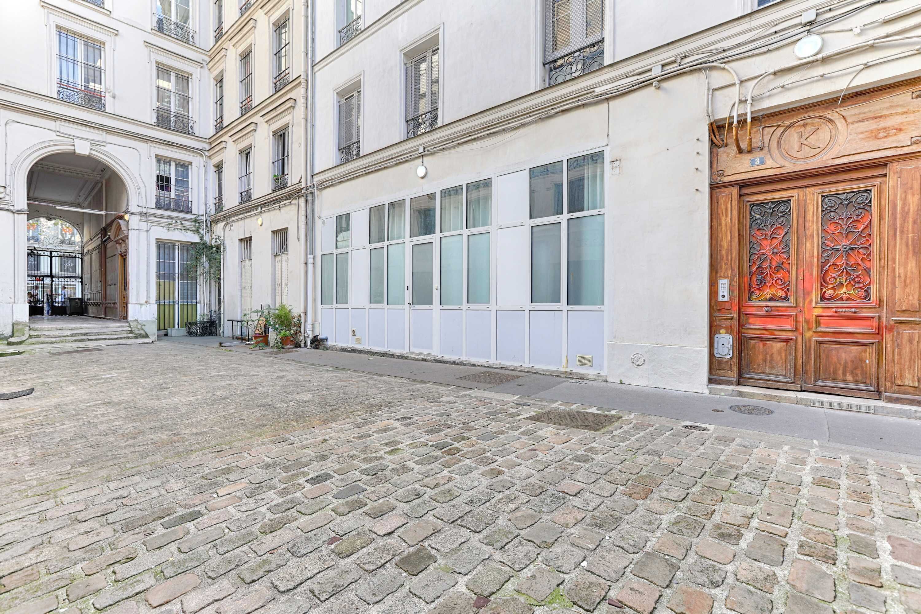 Un cortile con pavimento in pietra, una porta in legno con decorazioni in metallo elaborate e un edificio con ampie finestre e un balcone.