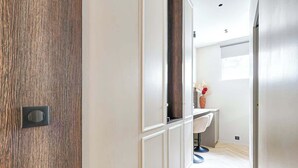 Een gang met een houten deur, een witte muur en een badkamer met een wit wastafelmeubel en een stoel.