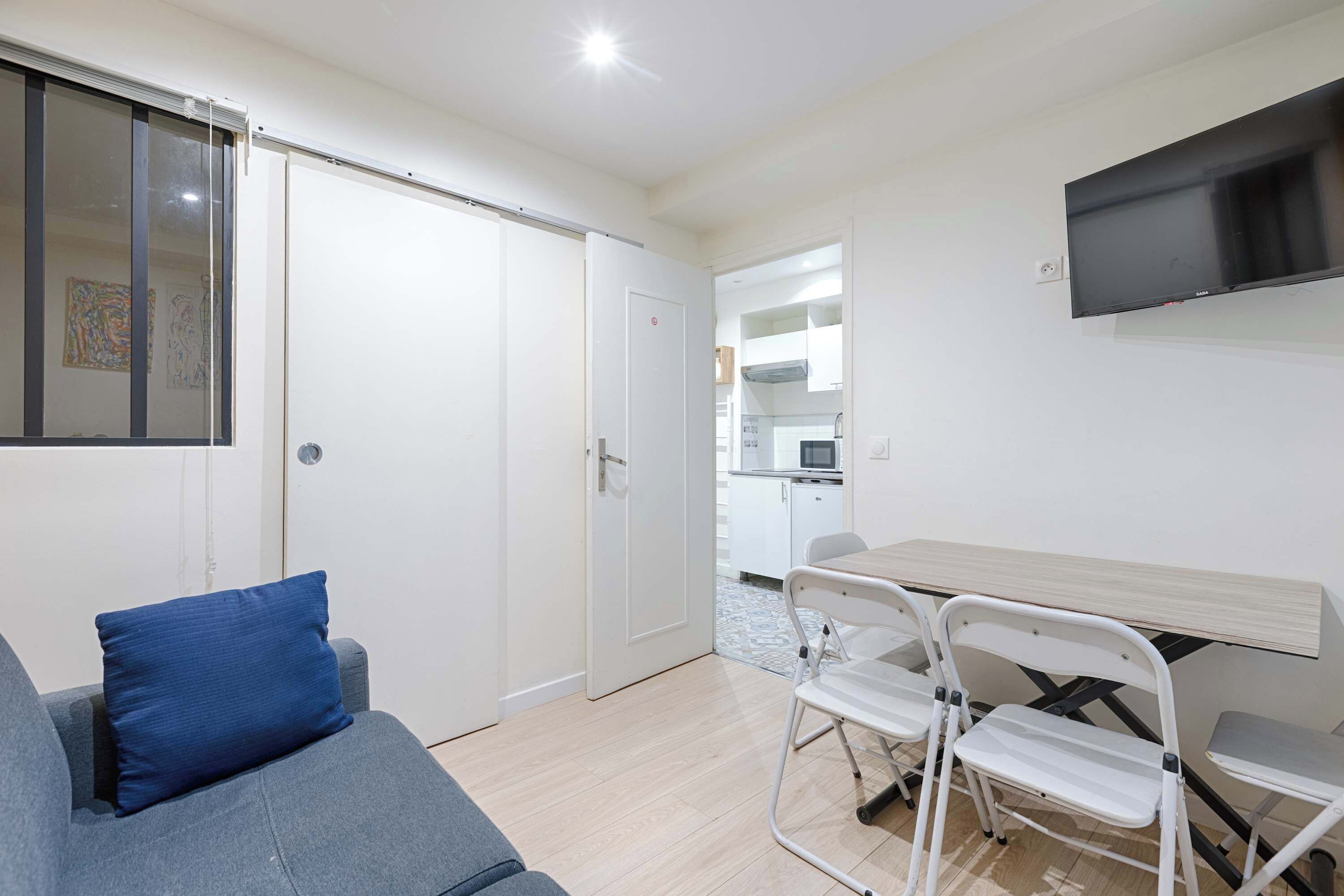 A modern room with a sofa, a dining table with chairs, a TV mounted on the wall, and a kitchen area in the background.