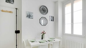 A dining area with a white table set for four, surrounded by white chairs, a wall clock, and framed pictures. - Appartement de charme cosy au cœur de Batignolles (Paris)
