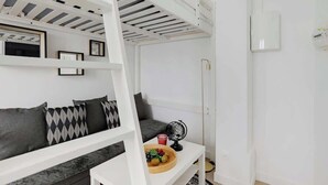 A modern, compact living space with a loft bed, a sofa with patterned cushions, a small table with a fruit bowl, and a globe. - Appartement de charme cosy au cœur de Batignolles (Paris)