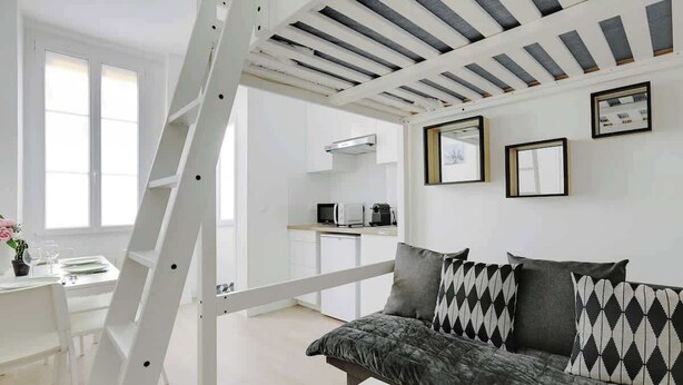 A modern, well-lit living room with a sofa, a dining area with a table and chairs, a kitchen area, and a white staircase. - Appartement de charme cosy au cœur de Batignolles (Paris)