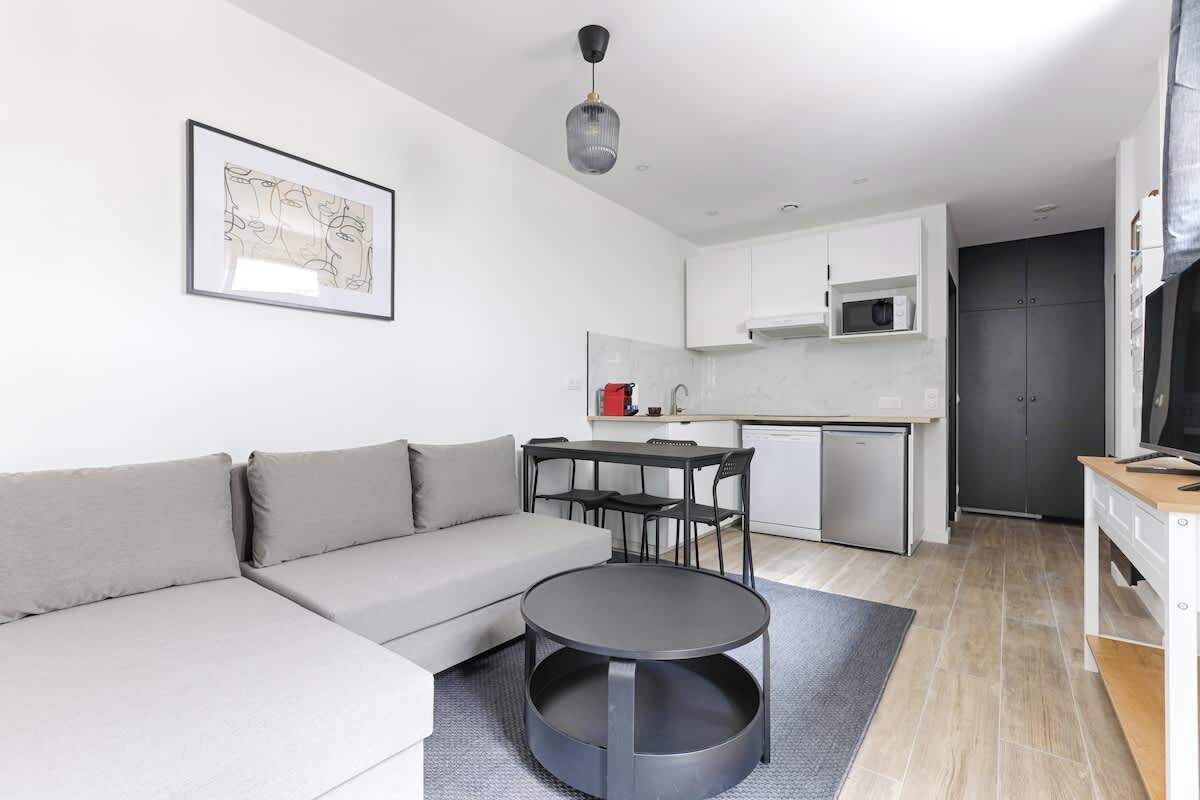 A modern living room with a grey sofa, a round black coffee table, and a dining area with a black table and chairs. There is a kitchen area with white cabinets and a microwave.
