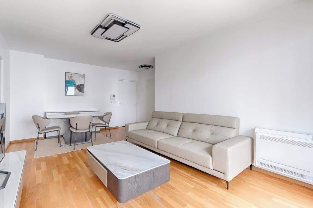 Una sala moderna con un sofá beige, una pequeña mesa de comedor con sillas, una mesita de centro de mármol y un radiador blanco.