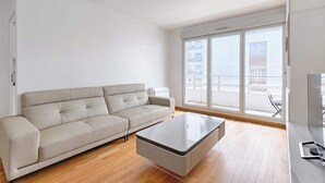 Un salon moderne avec un canapé et une table basse en verre, de grandes fenêtres offrant une vue sur les immeubles et un balcon.