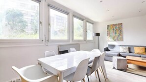 Un salón moderno con una mesa blanca, sillas y un sofá gris. Cuenta con ventanas amplias con persianas, un radiador y un cuadro abstracto y colorido en la pared.
