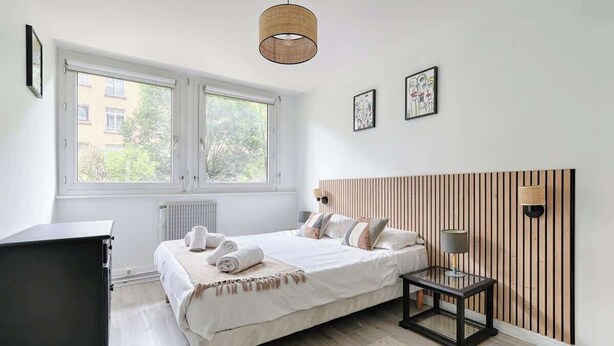 Un dormitorio con cama, mesita de noche, lámpara y ventana con vistas a edificios.