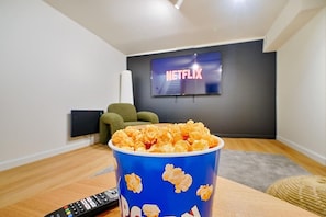 Una habitación con una pantalla grande mostrando el logotipo de Netflix, un cubo azul de palomitas de maíz y un control remoto sobre una mesa de madera.