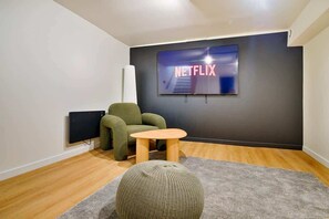 A modern room with a flat-screen TV displaying 'NETFLIX', a green armchair, a small wooden table, and a knitted pouf on a grey rug.