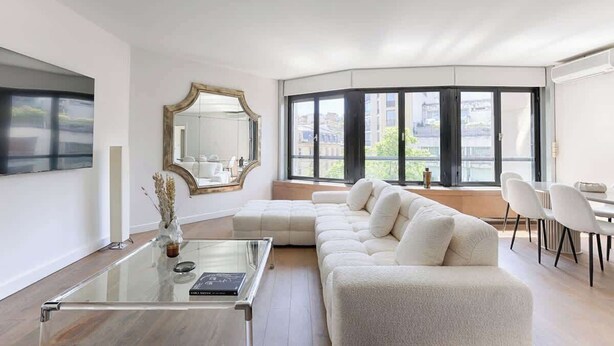 A modern living room with a glass coffee table, a white sofa, a large mirror, and a dining area with chairs.