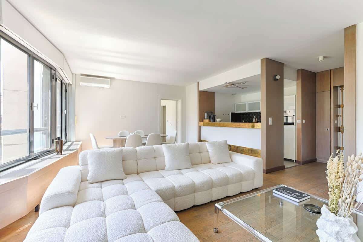 A modern living room with a sectional sofa, a glass coffee table, and a view of the kitchen through an open doorway.