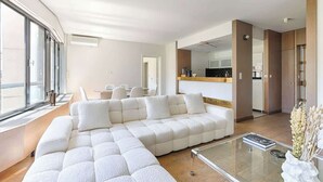 A modern living room with a sectional sofa, a glass coffee table, and a view of the kitchen through an open doorway.
