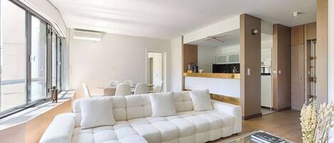 A modern living room with a sectional sofa, a glass coffee table, and a view of the kitchen through an open doorway.