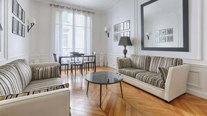 A living room with a sofa, armchair, and a round coffee table. There is a dining table with chairs and a lamp. - Appartement de Luxe sur les Invalides (Paris)