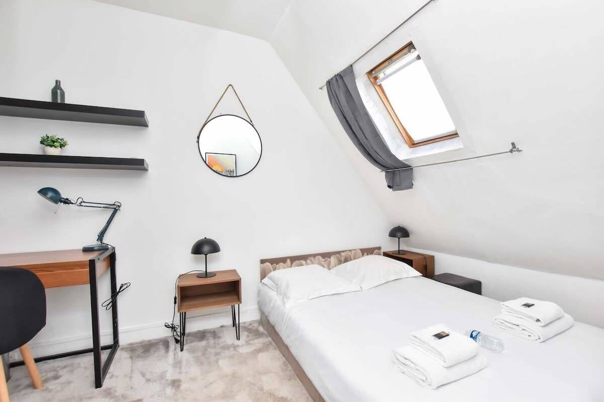 A modern hotel room with a desk, a bed, a bedside table with a lamp, a mirror, and a window with curtains.