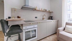 Una cocina compacta con horno integrado, un lavamanos y una estufa. En la zona de estar contigua hay una silla y un sofá.