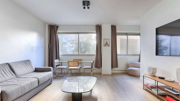 Un salon moderne avec un canapé gris, une table basse, une table à manger avec des chaises et une grande fenêtre avec des rideaux.