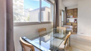 Un salon moderne avec une table à manger en verre, deux fauteuils en osier, une bibliothèque et une vue sur les immeubles à travers de grandes fenêtres.
