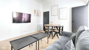 Un salon moderne avec un canapé gris, une petite table, un téléviseur fixé au mur et un coin repas comprenant des chaises et une table.