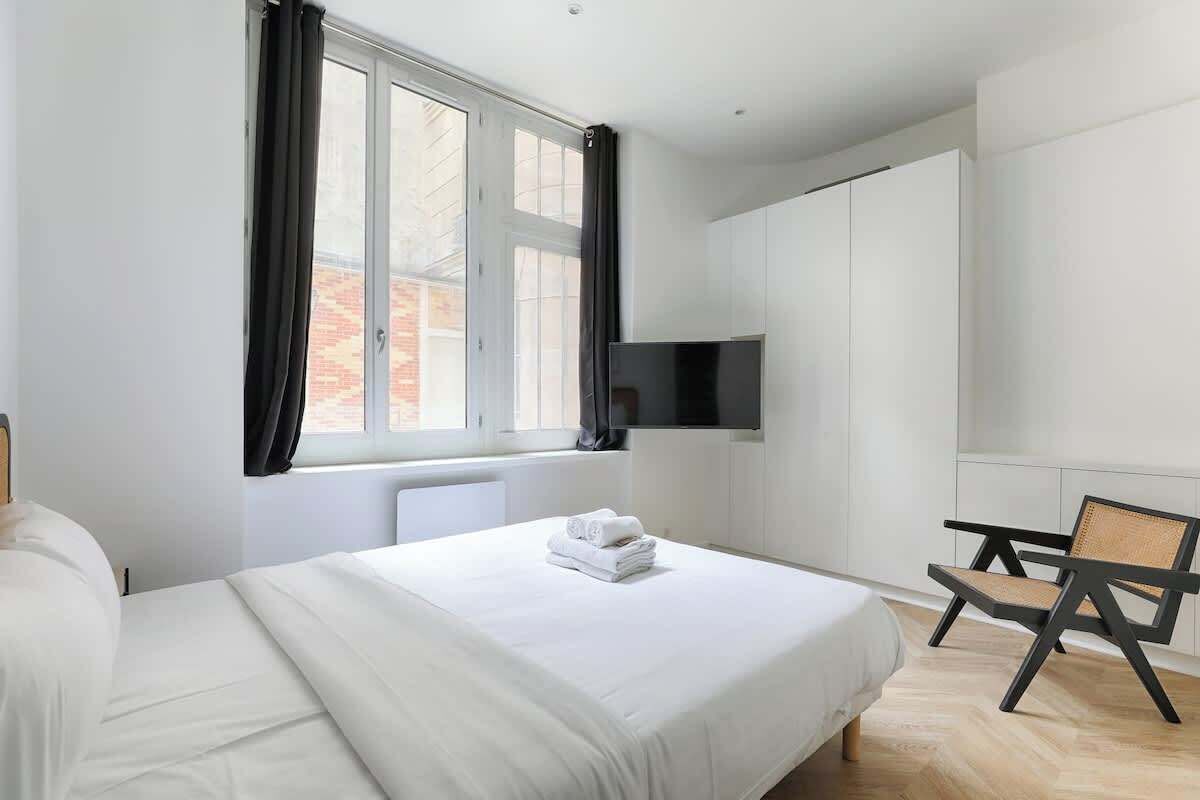 A modern hotel room with a large bed, a flat-screen TV mounted on the wall, and a window with curtains.
