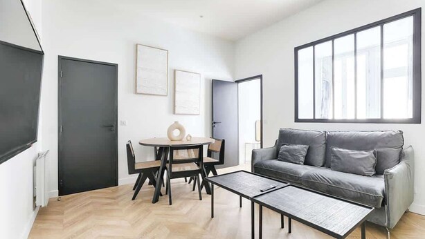 Un salon moderne avec un canapé gris, une table de salle à manger ronde entourée de chaises et une grande fenêtre laissant entrer la lumière naturelle.