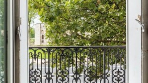 Ein offener Balkon mit schmiedeeisernem Geländer, freie Sicht auf einen Baum und ein Gebäude im Hintergrund.
