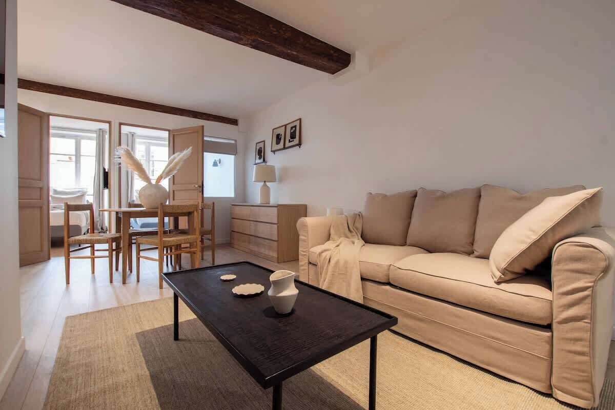 A living room with a sofa, coffee table, and chairs. There is a window with curtains and a lamp on a side table.