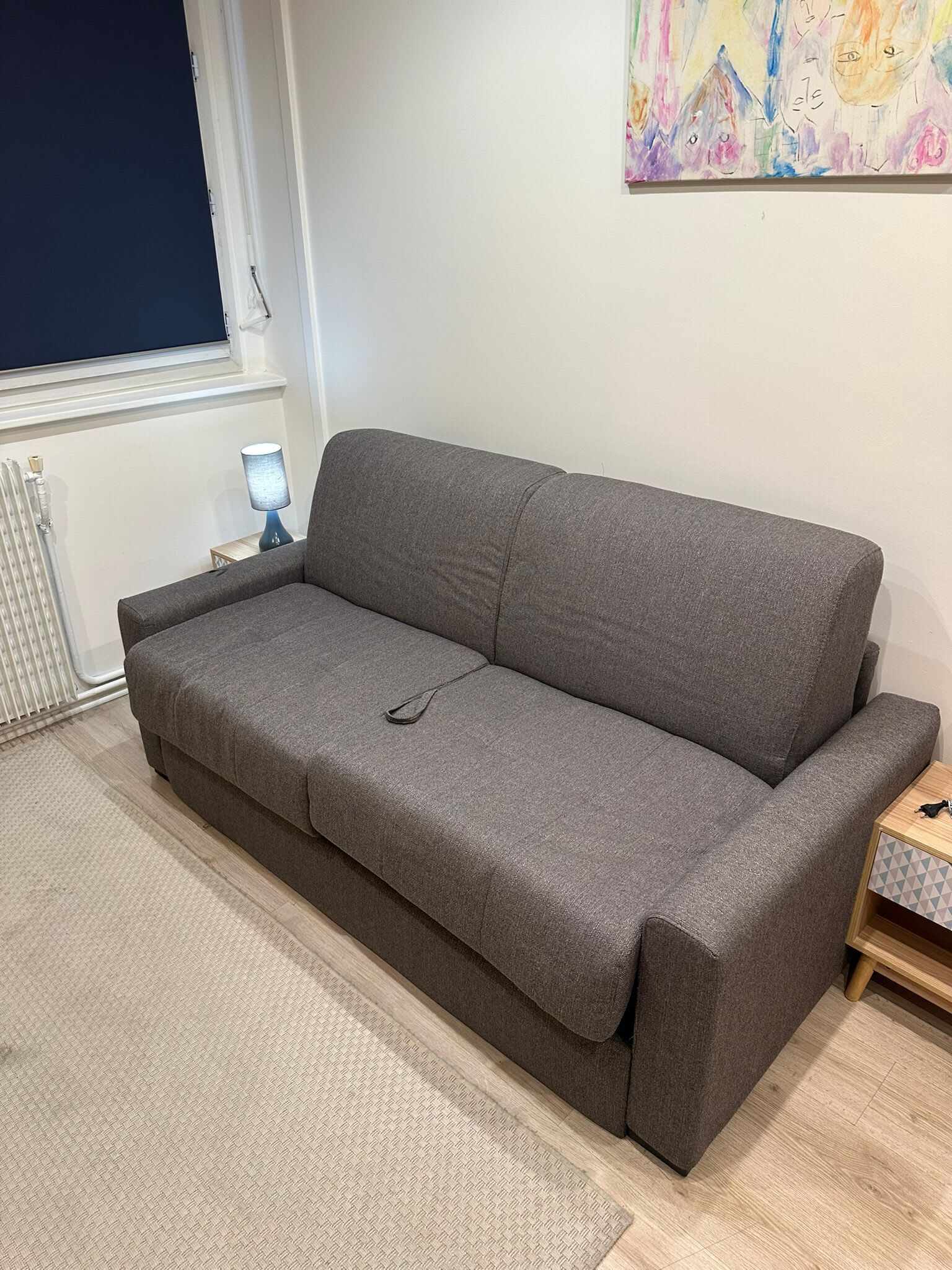 Un canapé gris dans une pièce équipée d’un radiateur, d’une lampe et d’une petite table d’appoint en bois.