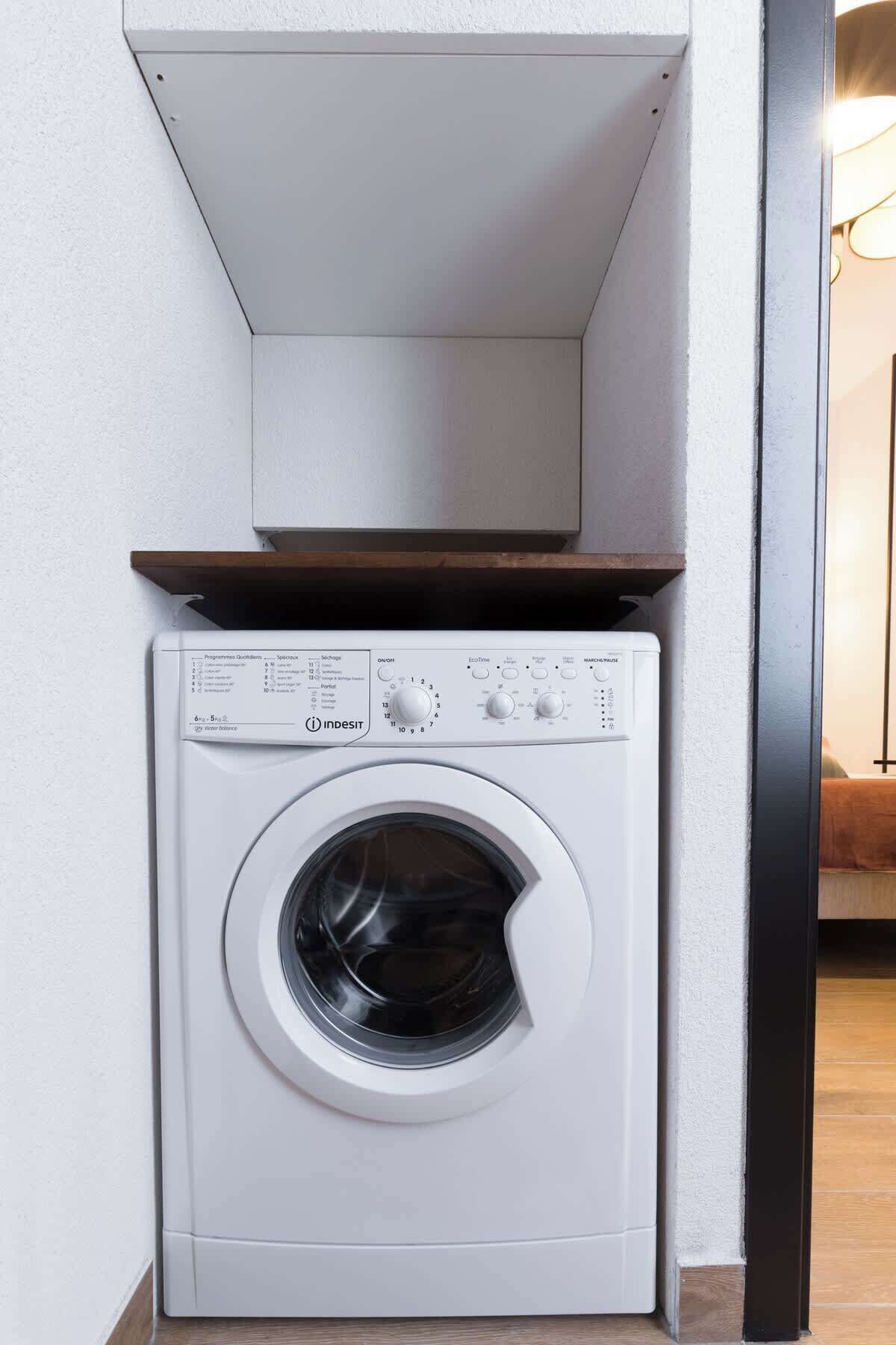 A compact, white front-loading washing machine with a control panel on top, installed in a corner of a room with white walls and a wooden floor.