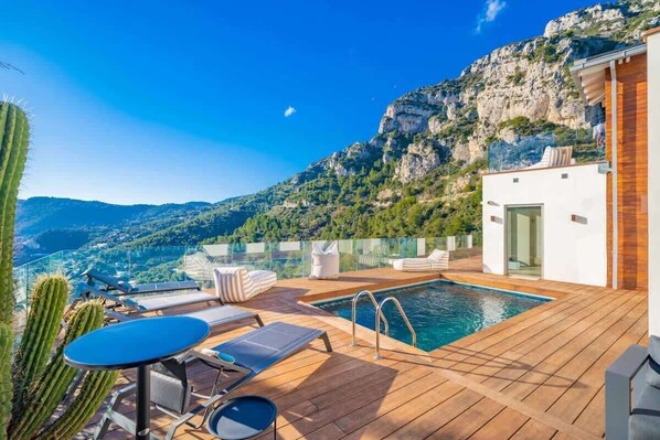 Un espace aménagé au bord de la piscine, avec vue sur les montagnes, une terrasse en bois et un bâtiment moderne.