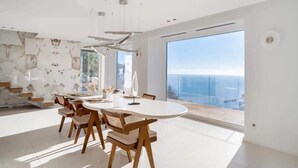 Et moderne spiseområde med et hvidt bord og stole, store vinduer og udsigt til havet.