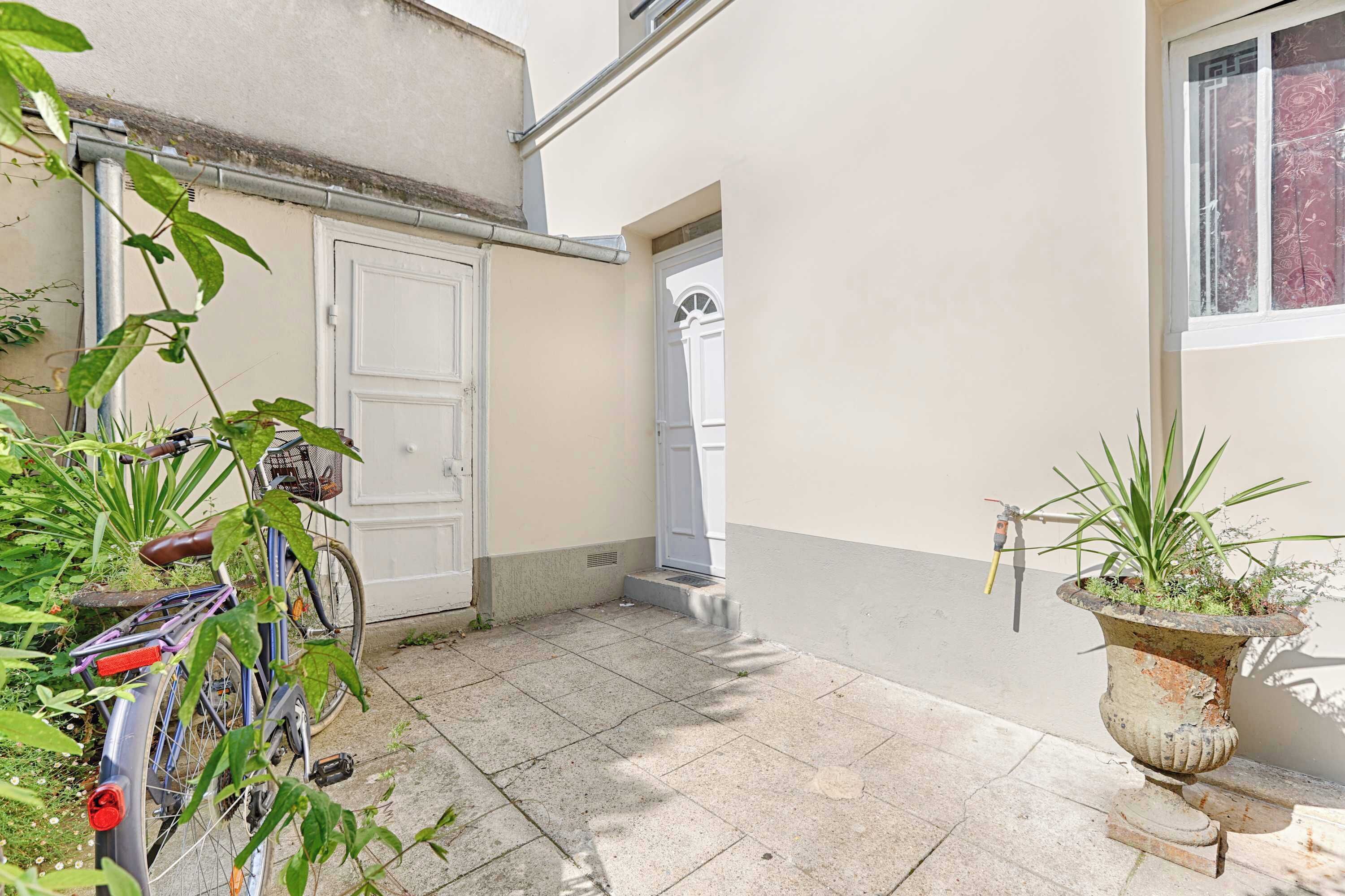 A narrow outdoor corridor with a bicycle parked on the left, a potted plant on the right, and a door at the end.