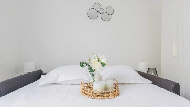 Uma cama bem arrumada com roupa branca, um cesto de tecido com velas e um vaso com flores.