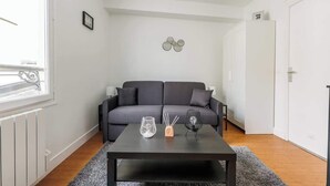Uma sala de estar moderna com um sofá cinzento, uma mesa de centro preta e um tapete cinzento. Há molduras de quadros na parede e um radiador.