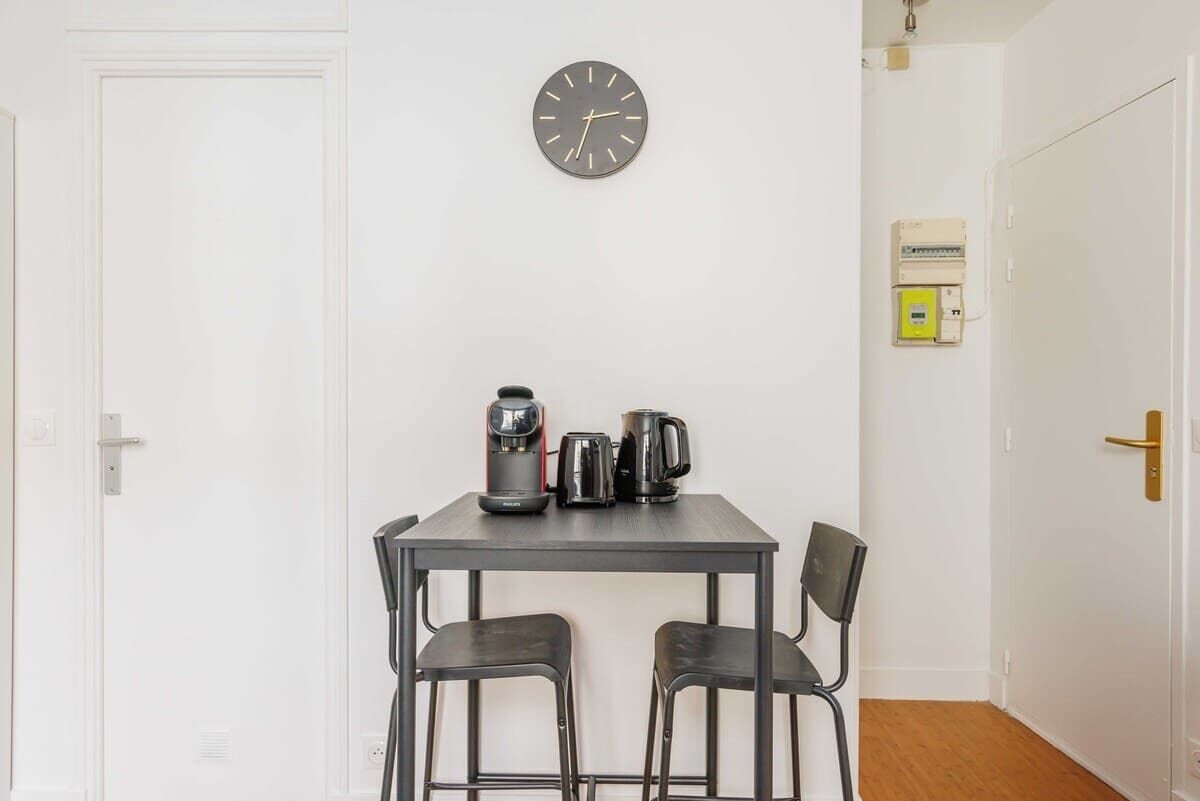 A small room with a white door, a wall clock, a table with two chairs, and a coffee maker.