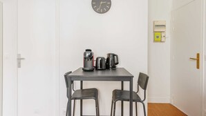 Um quarto pequeno com porta branca, um relógio de parede, uma mesa com duas cadeiras e uma cafeteira.