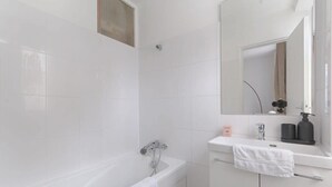 A modern bathroom with white tiles, a bathtub, a sink with a mirror, and a towel rack. - Laugier Ternes : Appartement Cosy en Bail Mobilité (Paris)