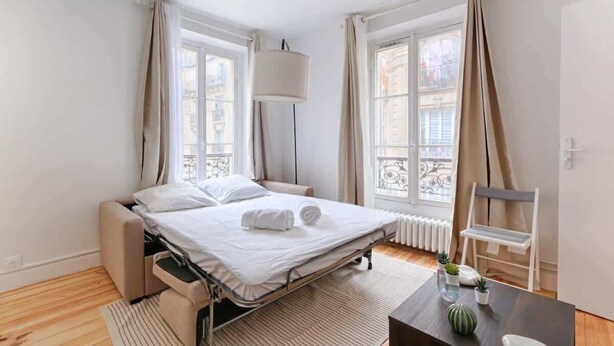 A modern hotel room with a bed, a bedside table, a chair, and a view of the city through large windows. - Laugier Ternes : Appartement Cosy en Bail Mobilité (Paris)