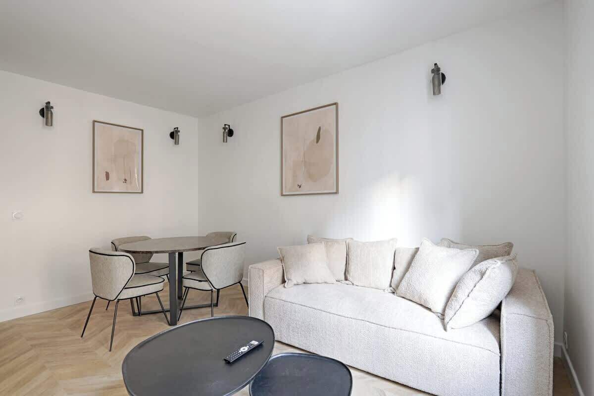 Un salon moderne comprenant un canapé, une table basse et une table à manger avec des chaises. On y trouve deux luminaires fixés au mur et deux œuvres d’art encadrées.