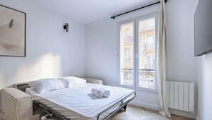 Een minimalistische slaapkamer met een bed, een radiator en een raam met uitzicht op gebouwen.