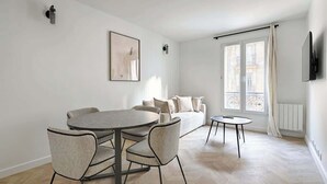 Een moderne woonkamer met een ronde eettafel, twee stoelen, een bank en een klein bijzettafeltje. Er is een groot raam met gordijnen, een wandgemonteerde televisie en een ingelijst schilderij aan de muur.
