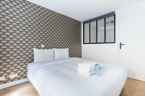 A hotel room with a patterned wallpaper, a bed with white linens, a window with blinds, and a door.
