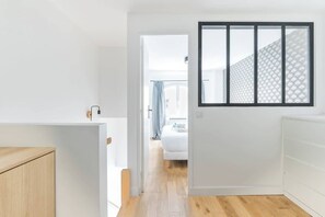 A hallway with a wooden floor, a white wall, and a window with a geometric pattern.