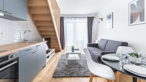 A modern kitchen with grey cabinets, a wooden staircase, a dining area with a grey rug, and a grey sofa. - Marivaux/Opéra : Charming & Cosy Duplex 4P-1BR (Paris)
