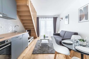 A modern kitchen with grey cabinets, a wooden staircase, a dining area with a grey rug, and a grey sofa.